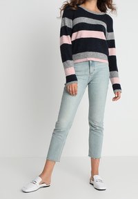 Žena v námořnickém, šedém a růžovém pruhovaném svetr, světle modrých zkrácených džínách a bílých slip-on mokasínech stojí na bílém pozadí.