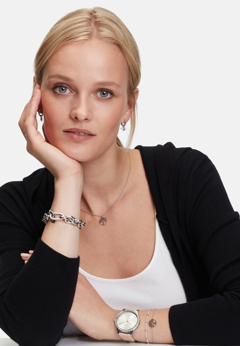 Silberner Schmuck mit einem Kettenarmband, einer Baum-Anhänger-Halskette und einer Uhr mit rundem Ziffernblatt; Modell trägt eine schwarze Strickjacke und ein weißes Oberteil.