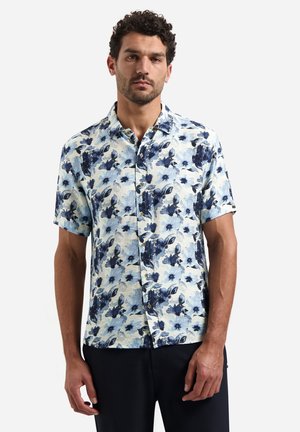 Mann trägt ein kurzärmeliges Hemd mit einem blauen Blumenmuster und dunkle Hosen, steht vor einem schlichten Hintergrund.