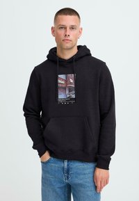 Sudadera de algodón negra con un bolsillo frontal, que presenta un estampado gráfico colorido en el pecho. Modelo vistiendo jeans azules, de pie contra un fondo claro.