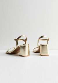 Sandalias abiertas de dedo en metal dorado con tacón bloque, que presentan correas ajustables en el tobillo y una superficie suave y brillante.