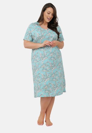 Kobieta z długimi ciemnymi włosami, ubrana w jasnoniebieską sukienkę w kwiaty do kolan, stojąca boso na jednolitym tle, uśmiechająca się i patrząca w dół.