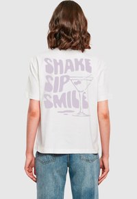 White cotton t-shirt featuring a lavender graphic design on the back with the text "SHAKE SIP SMILE" and a martini glass illustration.