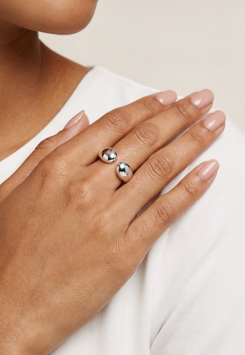Main avec des ongles polis portant une bague ouverte argentée brillante avec deux extrémités arrondies, reposant sur une chemise blanche près de l'épaule.