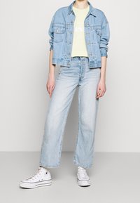 Chaqueta de mezclilla azul claro, camiseta gráfica verde pálido, pantalones vaqueros anchos azul claro y zapatillas altas blancas. Diseño sencillo y estilo informal.