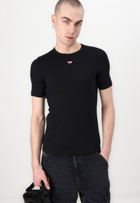 Svart tettsittende t-skjorte med korte ermer laget av glatt stoff, med en liten rød logo på brystet, kombinert med mørke cargo-shorts.