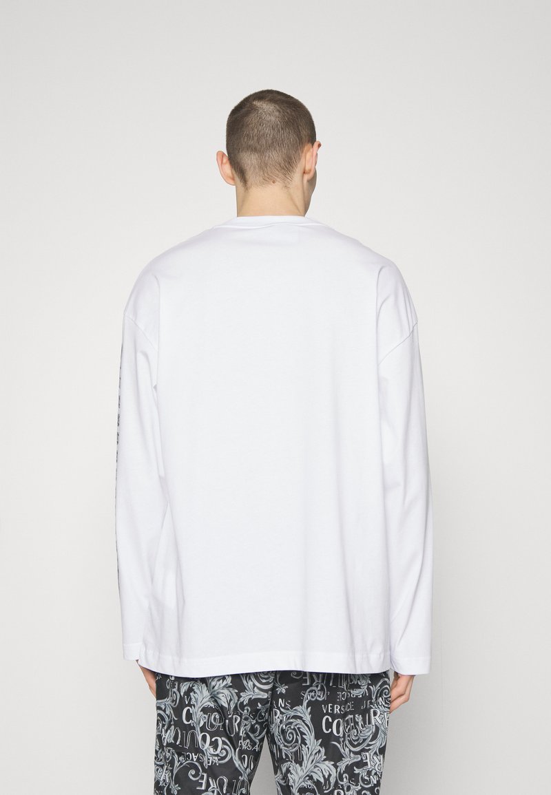 Camiseta blanca de manga larga con cuello redondo, corte relajado y hombros caídos; combinada con pantalones cortos negros estampados con texto y remolinos.