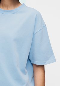 Άτομο που φοράει ένα ανοιχτό μπλε κοντομάνικο μπλουζάκι, εμφανίζοντας τον άν�ω κορμό και το δεξί μπράτσο σε έναν απλό λευκό φόντο.