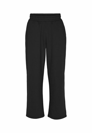 Personne portant un pantalon de survêtement noir ample et des baskets beiges, debout les mains dans les poches devant un fond uni.