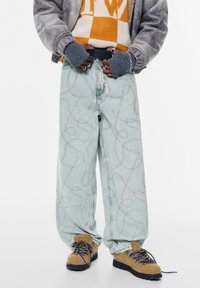 Jeans palatale de culoare albastru deschis, cu un model de cusătură în bucle, asortați cu pantofi texturați maro și o jachetă gri purtată peste un pulover portocaliu și alb.