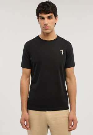 Mand iført en sort t-shirt med korte ærmer med et lille beige logo på venstre bryst og beige bukser, stående foran en ensfarvet baggrund.