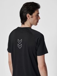 Ung mann med mørkt hår iført en svart kortermet skjorte med to hvite nedadgående Chevron-piler på øvre del av ryggen, vendt mot høyre.