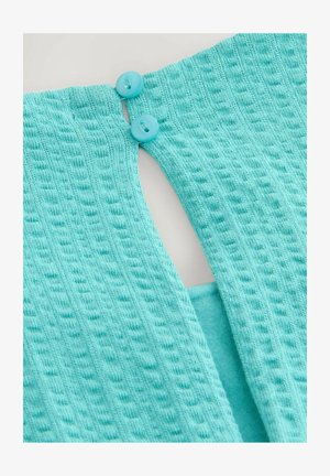 Türkiissinine kampsun, millel on tekstuuriga kudumine, kaks sinist nööpi taga ning avatud seljaga disain, millel on ristkülikukujuline lõikeauk.