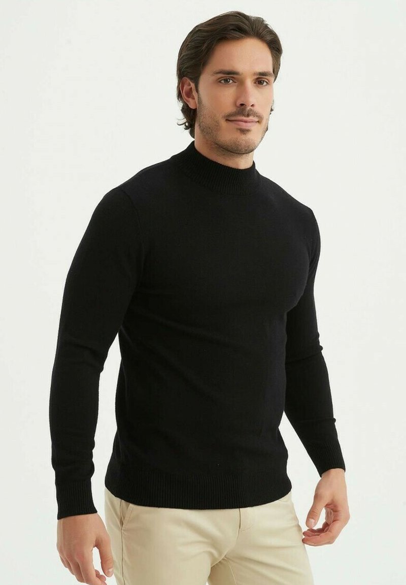 Pull à col montant ajusté noir en tissu doux, avec des manches longues et des poignets côtelés, associé à un pantalon beige clair.