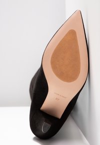 Chaussure à talon aiguille noire à bout pointu, inclinée pour montrer la semelle beige lisse avec la taille 37 et l'inscription "Fabriqué en Italie" en relief.