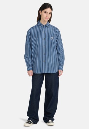 Frau trägt ein blaues, vertikal gestreiftes Hemd mit Knöpfen, dunkle Jeans mit weitem Bein und schwarz-weiße Sneakers, steht vor einem einfarbigen Hintergrund.