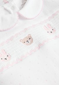 Vêtement en coton blanc avec un col en forme de coquille, orné d'une broderie rose d'un ours et de deux lapins sur une bande froncée. Pois roses sur l'ensemble.