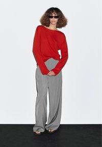 Pull rouge oversize associé à un pantalon large gris clair. Le modèle porte des lunettes de soleil noires et des sandales, posant devant un fond blanc.