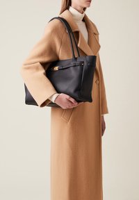 Black leather tote bag with a textured finish, gold-tone hardware, and dual elongated handles, held next to a camel-colored coat.