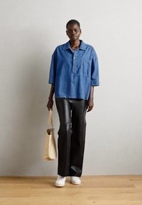 Denimhemd mit Knopfleiste, kurzen Ärmeln. Schwarze weite Hosen aus Kunstleder. Beiger Shopper, weiße Sneakers. Einfarbiger Hintergrund.