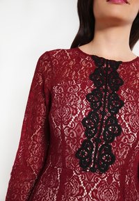 Burgundy spetsklänning har ett blommönster, långa ärmar och en svartsbroderad detalj nedanför i mitten med rund detaljer.