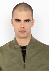 Chaqueta bomber color verde oliva con cuello acanalado, cremallera frontal y logo sutil. La tela tiene una textura suave y presenta una silueta ajustada.