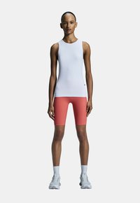 Witte mouwloze top en koraalkleurige shorts met een aansluitend ontwerp. Model in lichtgrijze sportschoenen en witte sokken, staand op een effen achtergrond.