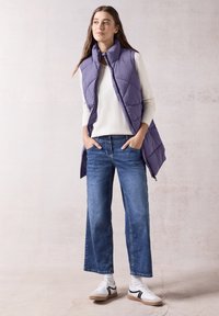 Hellblaue, puffy Weste über einem cremefarbenen Pullover, kombiniert mit weit geschnittenen blauen Jeans und weißen Turnschuhen mit schwarzen Akzenten.