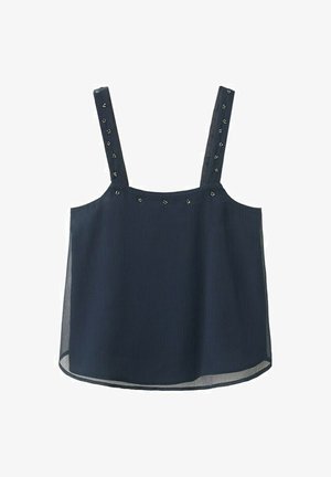 Jeune femme portant un haut sans manches noir et un pantalon à jambes larges, avec des talons argentés et des boucles d'oreilles pendantes, debout devant un fond beige uni.