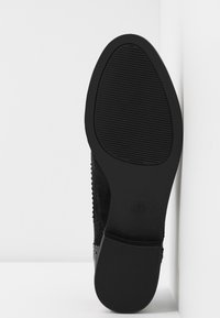 Semelle de chaussure noire avec une surface en caoutchouc texturée, présentant des rainures horizontales, une forme courbée et un léger talon, sur un fond blanc.