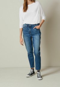Camisa blanca de manga larga ligera combinada con jeans de mezclilla azul de tiro alto, ligeramente descoloridos, y zapatillas negras de caña alta.