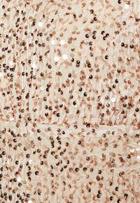 Tissu beige clair recouvert de sequins rose doré densément regroupés, avec une surface brillante et texturée, créant une apparence étincelante.