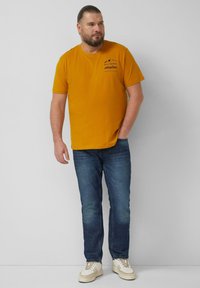 Mustard yellow cotton t-shirt with short sleeves, featuring a black graphic design. Paired with dark blue denim jeans and white sneakers.