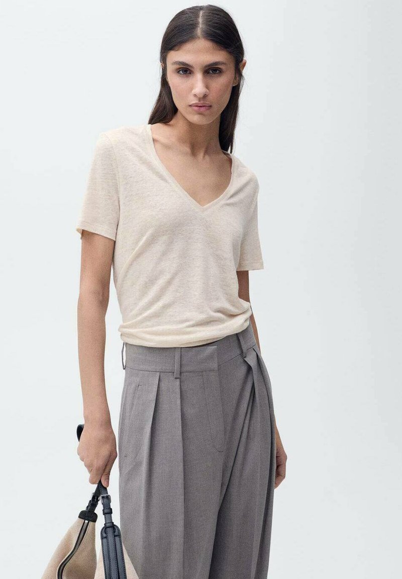 T-shirt beige à col en V, manches courtes, en tissu léger, associé à un pantalon gris taille haute plissé. Le mannequin tient un sac dans une main.