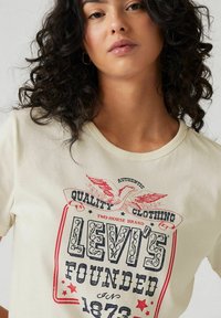 Crème-kleurige katoenen t-shirt met een grafisch ontwerp met "Levi's" in vet, sierlijk lettertype,accentueert met rode sterren en een adelaar.