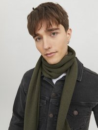 Sciarpa verde a coste realizzata in materiale morbido, drappeggiata attorno al collo sopra una giacca di jeans nera con dettagli in bottoni e una camicia bianca sotto.