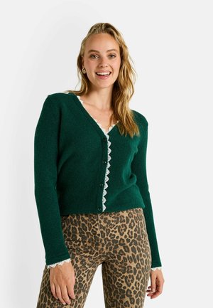 Cárdigan verde de punto con escote en V, con un borde de encaje a lo largo del escote y los puños, combinado con pantalones de estampado de leopardo.