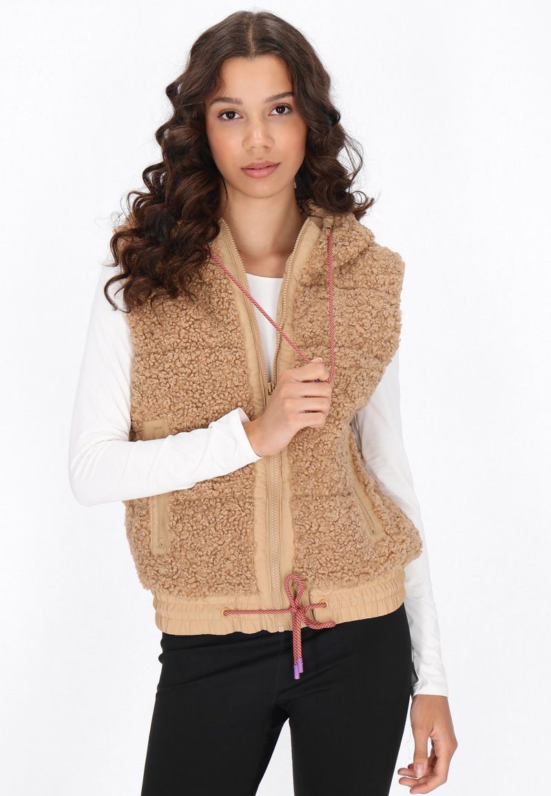 Beige Sherpa-Weste mit Reißverschluss, verstellbarem Kordelzugsaum und Seitentaschen. Getragen über einem weißen Langarmoberteil.