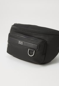 Black fabric pouch with a textured finish, featuring a top zip, a front zip pocket, and a silver metal loop accent. Logo displayed prominently.