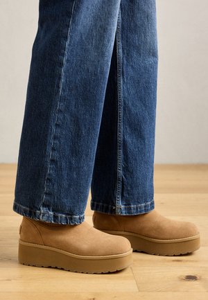 Brune suedesko med platå og en jevn tekstur, rund tå og elastisk ankel. Kombinert med vide denim-jeans. Bakgrunn med tregulv.