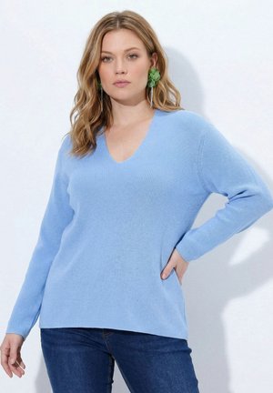 Femme aux cheveux châtain clair ondulés, portant des boucles d'oreilles fleurs vertes, un pull bleu clair à col en V et un jean bleu foncé, debout avec une main sur la hanche.