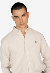 Camisa de cuadros clara en tonos beige, de material de algodón. Presenta un logo bordado azul en el pecho y un diseño de cuello clásico.