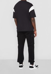 Camiseta de algodón negra con acentos blancos en los hombros, combinada con pantalones cargo negros que tienen bolsillos laterales y puños elásticos, usados con zapatillas negras y blancas.