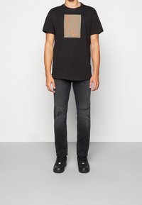 Czarna koszulka z bawełny z pomarańczowym logo na szarym kwadracie. Połączona z ciemnoszarymi dżinsami z przetarciami i czarnymi trampkami.