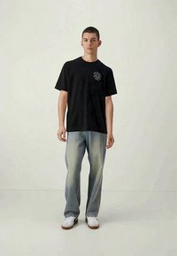 T-shirt de algodão preta com mangas curtas, detalhe gráfico no peito. Usada com jeans azul claro e tênis brancos, em pé sobre um fundo neutro.