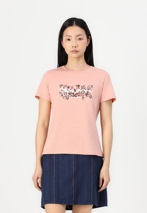 T-shirt in cotone rosa con maniche corte, caratterizzato da una stampa floreale in colori scuri sulla parte anteriore. Il modello lo indossa con una gonna a righe blu scuro.