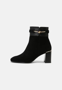 Bottines en daim noir avec un accent doré à l'avant, un talon brillant et un large revers noir. Elles présentent un élégant bout pointu et un intérieur rembourré.