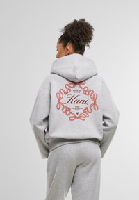 Sivý mikina s kapucňou, ktorá má okrúhly potlač s červenými stuhami a nápisom "Spread Love" a "Kani, The Brooklyn Way" na chrbte.