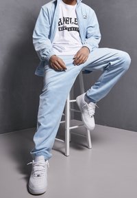 Světlomodrá tepláková souprava z měkké látky, obsahující bundu na zip a kalhoty se jemným vzorem, doplněná bílými teniskami.