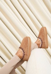 Pantofi platformă din piele întoarsă de culoare tabac cu o bandă cu imprimeu leopard, având o talpă texturată și un partea superioară căptușită. Așezați pe un fundal crem.